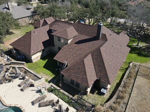 Brown-roofed house from a high angle. Green grass, pool, and trees surround the beige building.