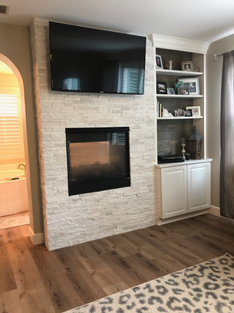 A room featuring a white stone fireplace with a mounted television above it and white built-in shelving beside it.