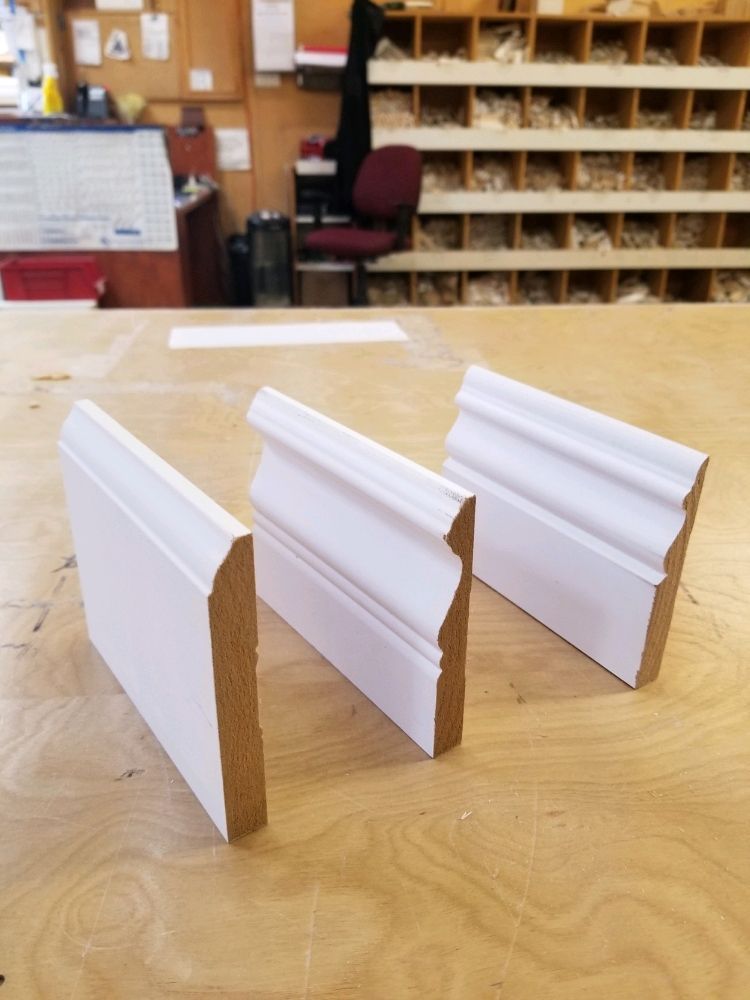 Three samples of white-painted baseboard molding with different profiles stand upright on a wooden workshop table.