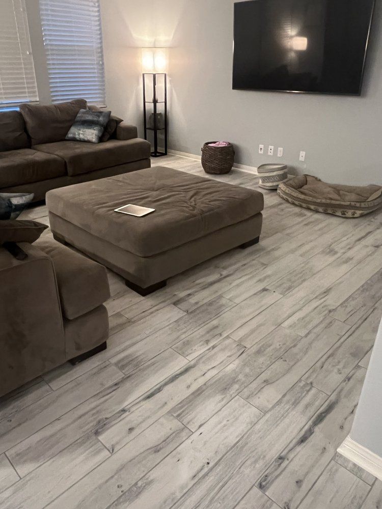 A modern living room with gray wood-look flooring, a large brown sectional sofa, an ottoman, a floor lamp, and a TV.
