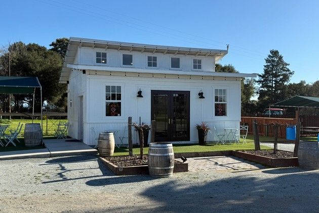 A small, white, single-story building with a clerestory roof and a central dark double door, surrounded by wine barrels.