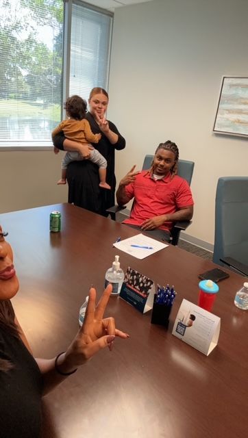 People at a table, signing documents. One person holds a child. Office setting, window in the background.