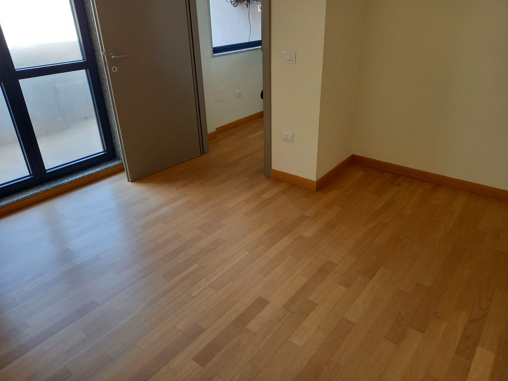 Stanza vuota con pavimento in legno chiaro, pareti beige e una porta vicino a una finestra.