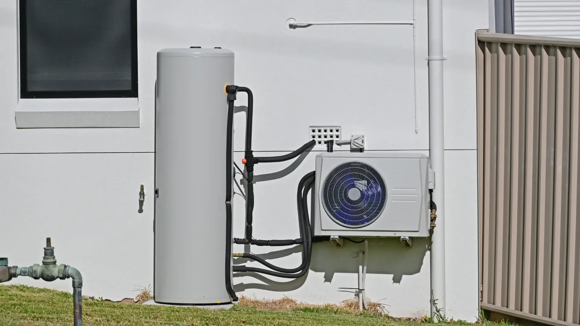 Outdoor utility equipment: gray tank, wall-mounted AC unit, pipes, and a beige fence beside a white building.