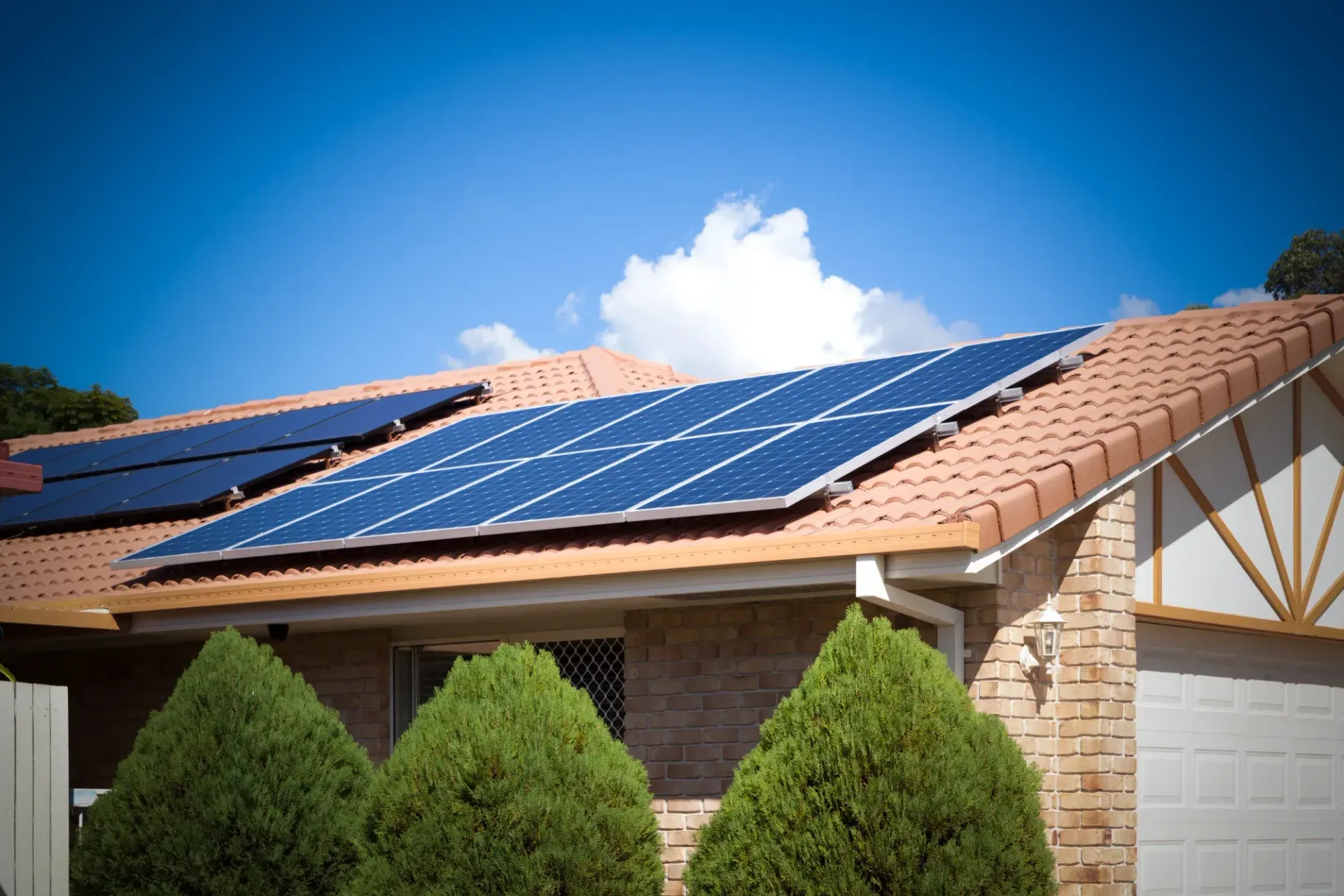 Solar Panels Installed On A Tan Tiled House Roof Under A Blue Sky, With Green Shrubs In Front — Macleay Energy Solutions In Kempsey, NSW