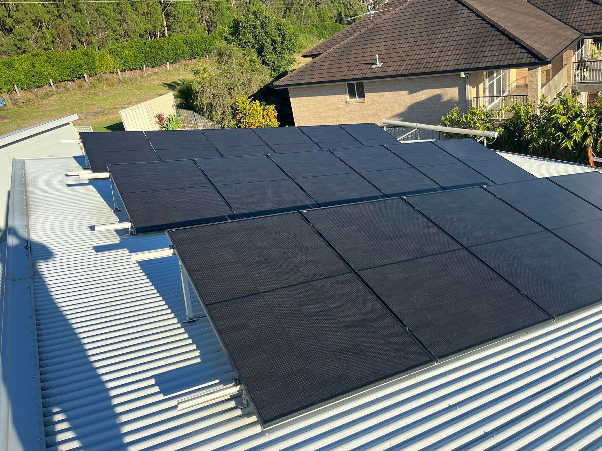 Solar panels mounted on a corrugated metal roof beside a house and trees — Macleay Energy Solutions In Crescent Head, NSW