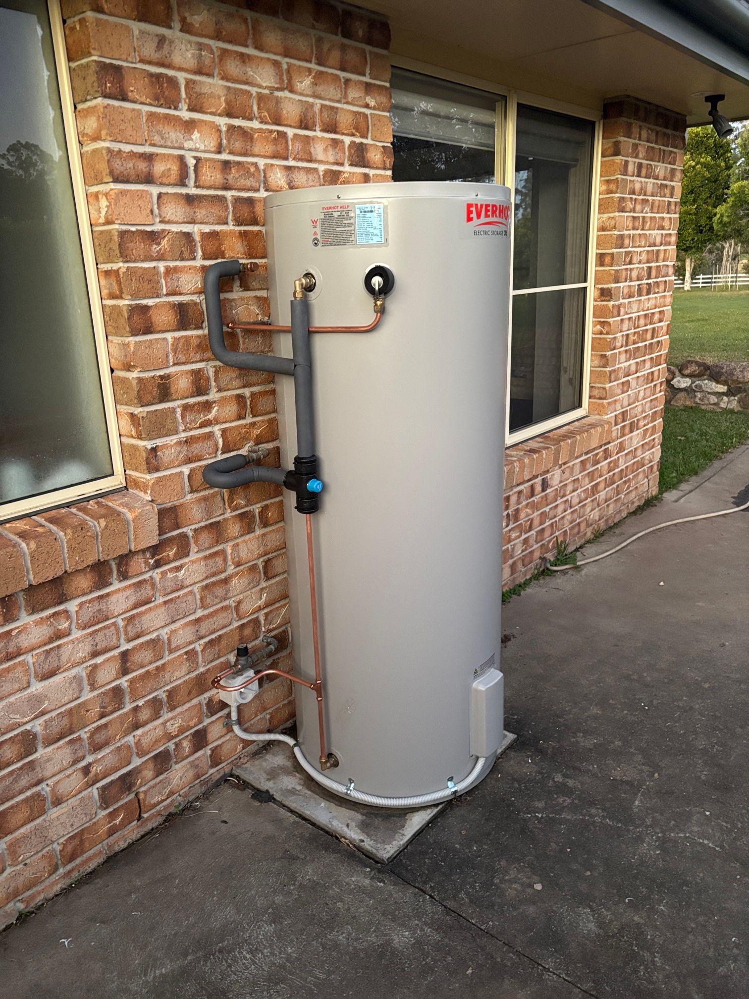 Tall White Cylindrical Water Heater Beside a Brick House Wall — Macleay Energy Solutions In Kempsey, NSW