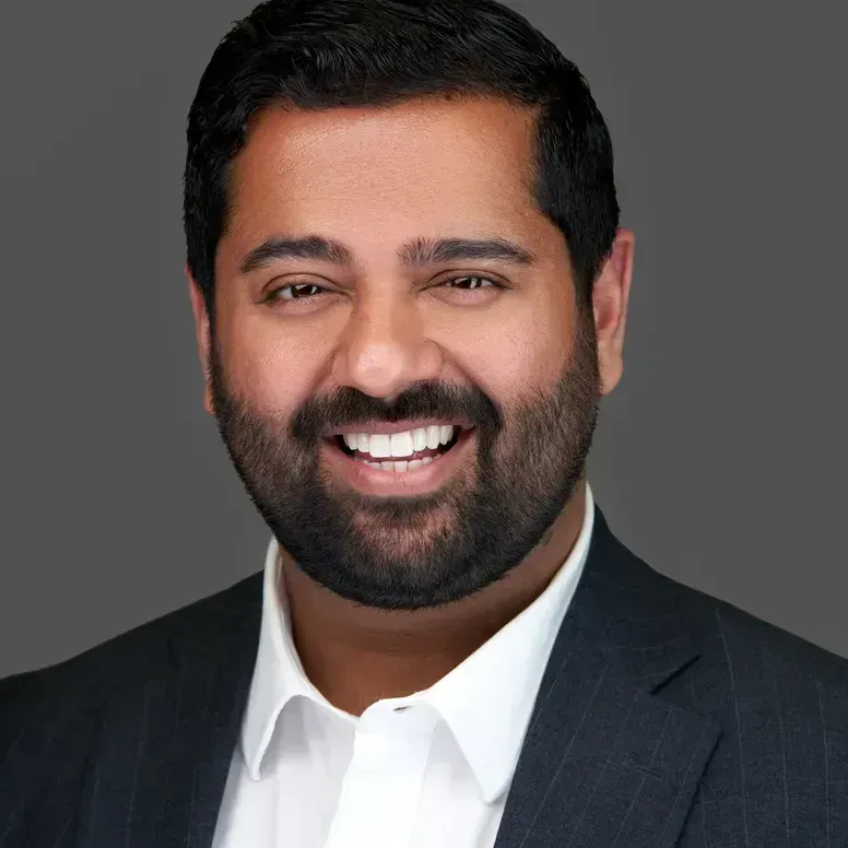 Man in suit smiling, with dark hair and a beard, against a gray background.