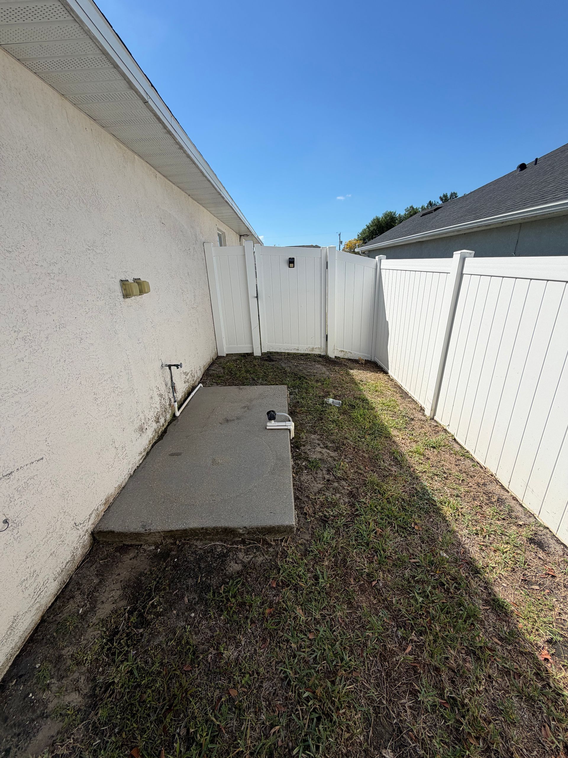 Narrow fenced side yard with white walls, gravel, a concrete slab, and a back gate under a blue sky