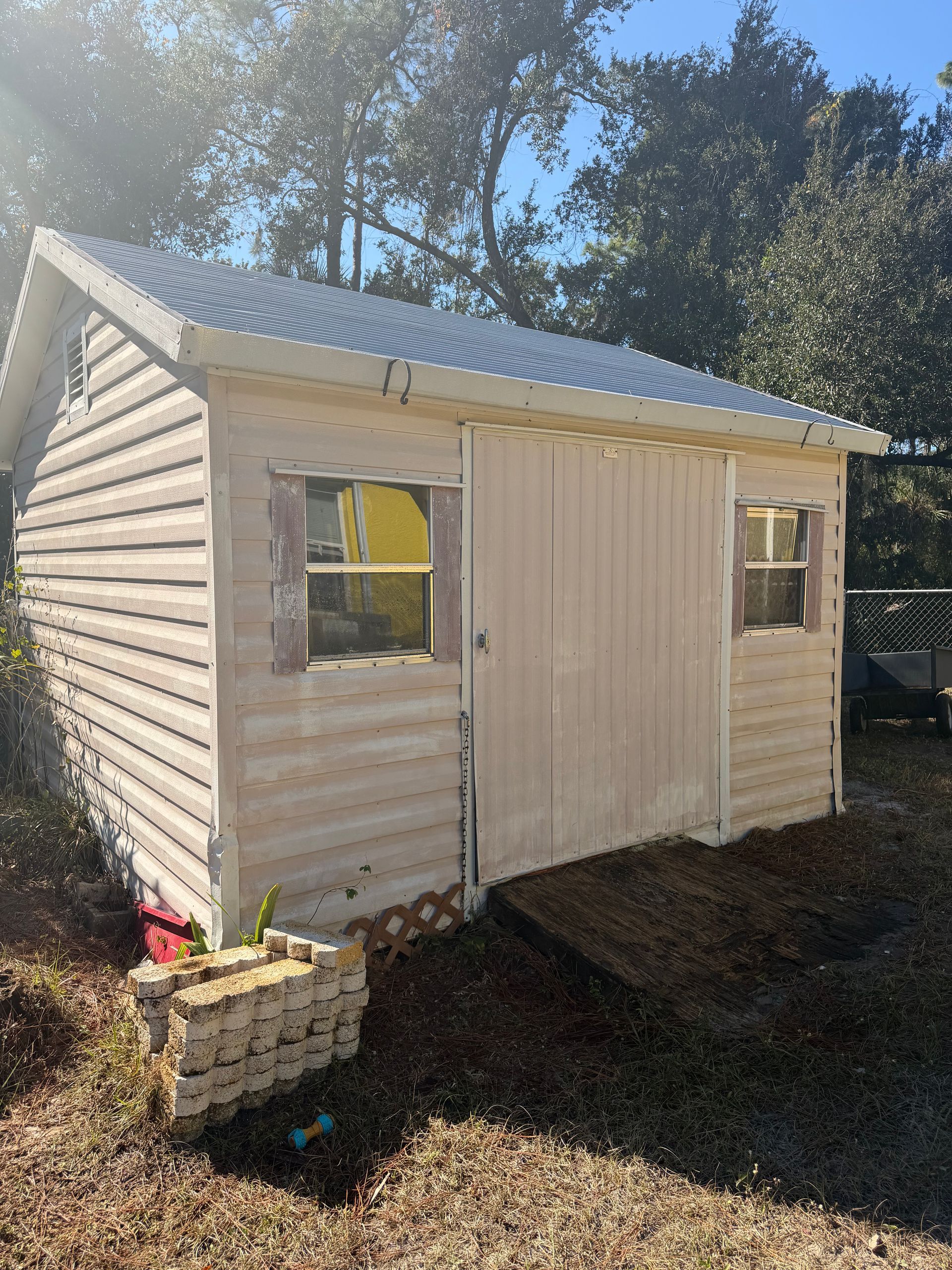 Small beige shed with corrugated metal roof in a sunny yard, with a window and double doors.