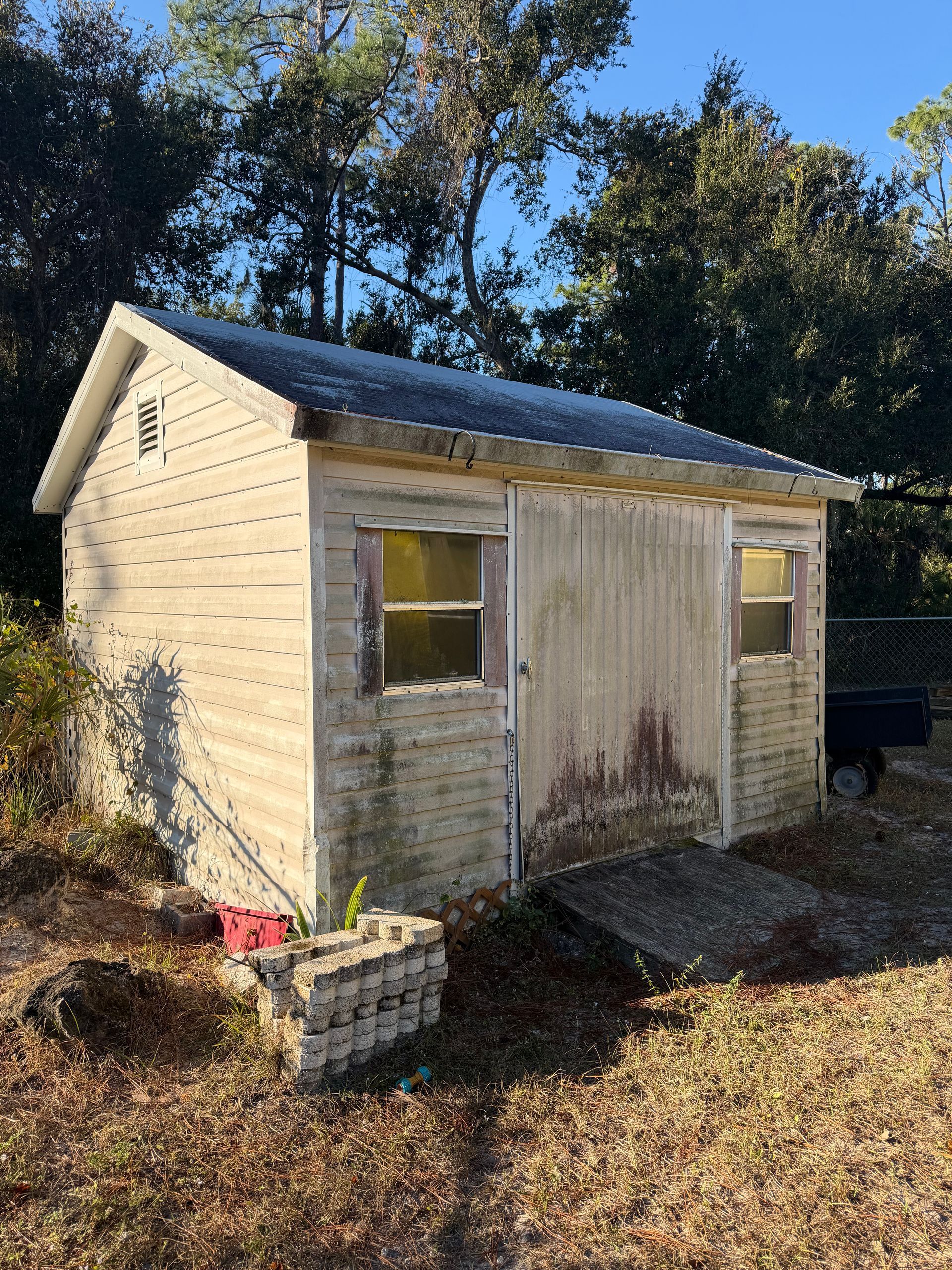 Small weathered shed with peeling paint and a corrugated roof in a grassy yard, with trees behind it