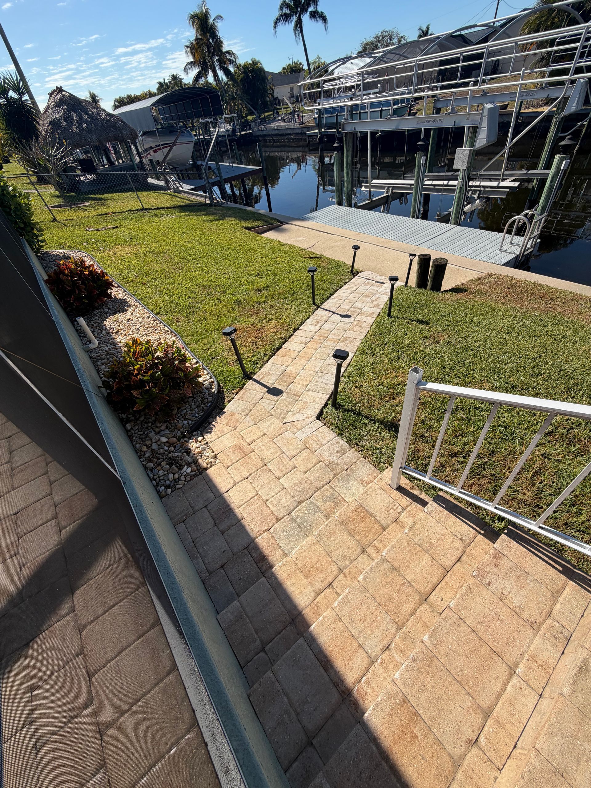 Sunlit backyard with stone path, green lawn, white fence, and waterfront dock in the background