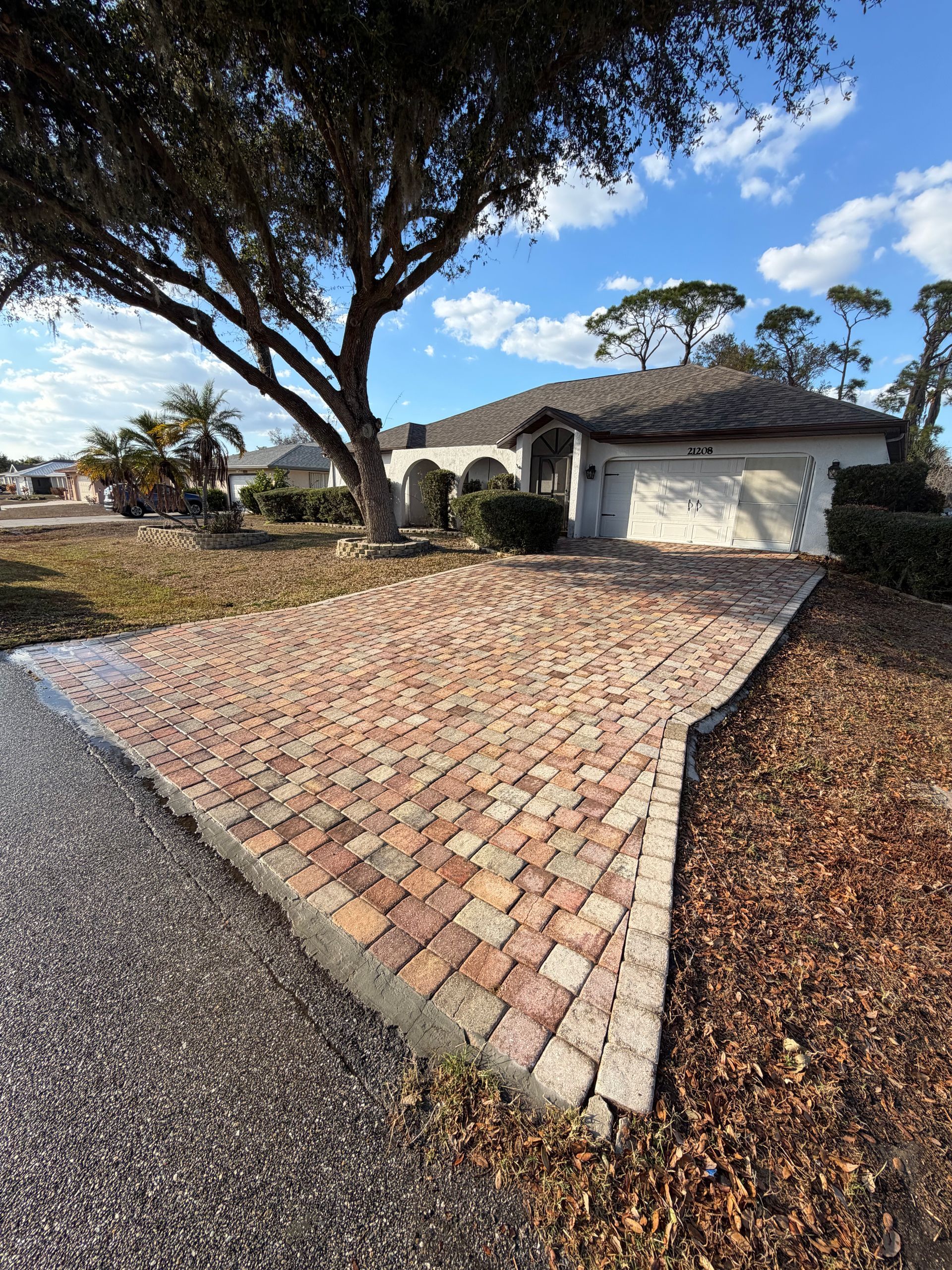 Brick driveway beside a white house with a large tree and gravel yard under a blue sky