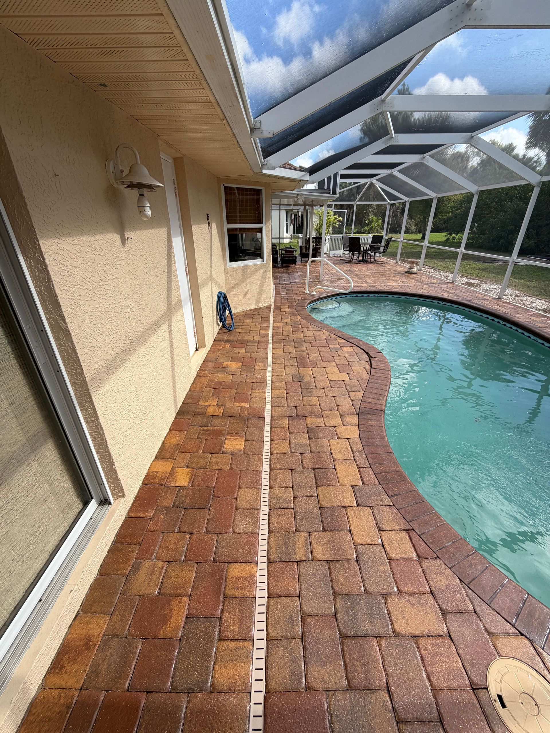 Covered pool deck with brick pavers beside a blue swimming pool and screened lanai under a cloudy sky