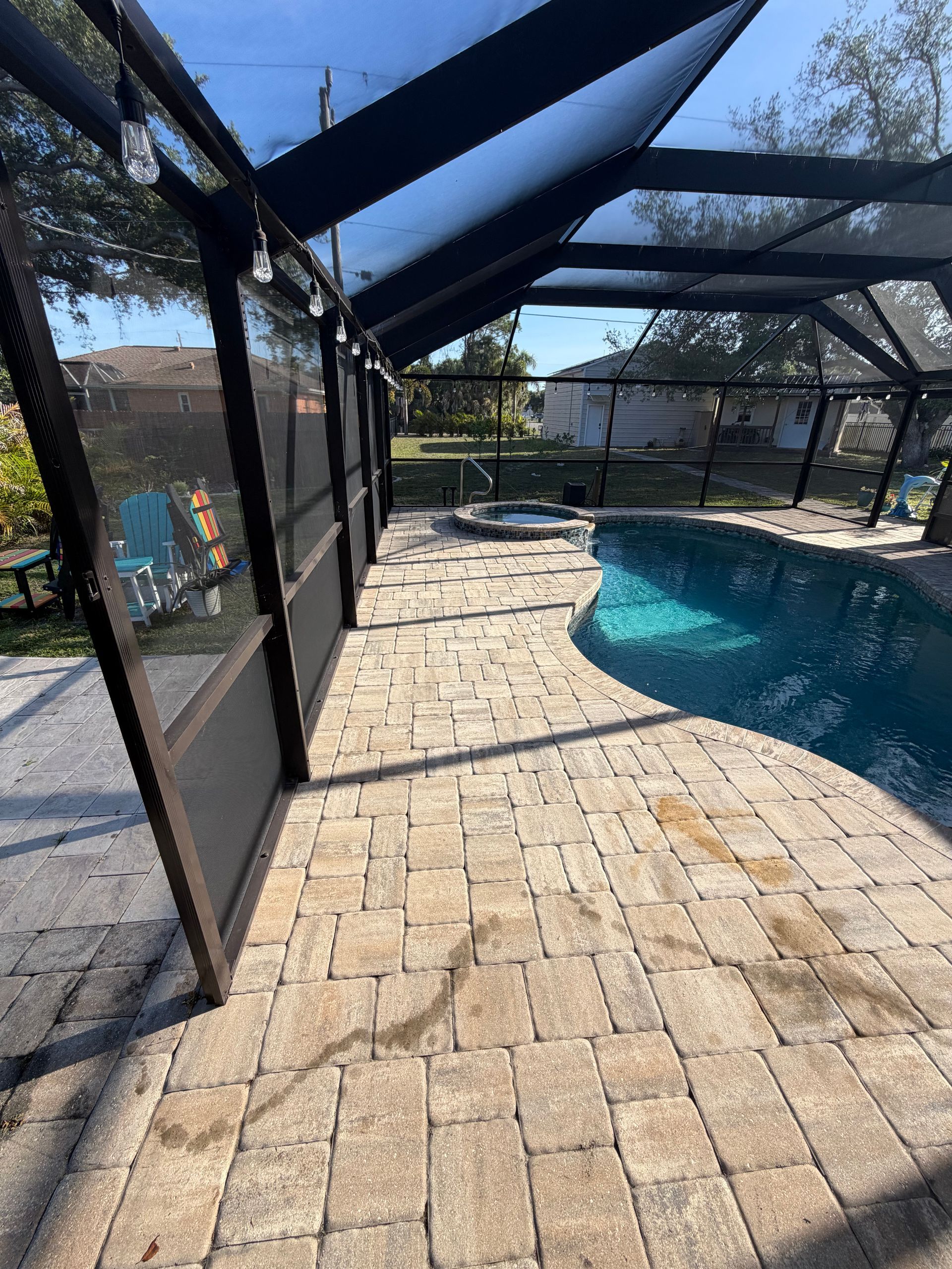 Screened poolside patio with brick pavers, black awning frames, and a blue swimming pool in bright sunlight