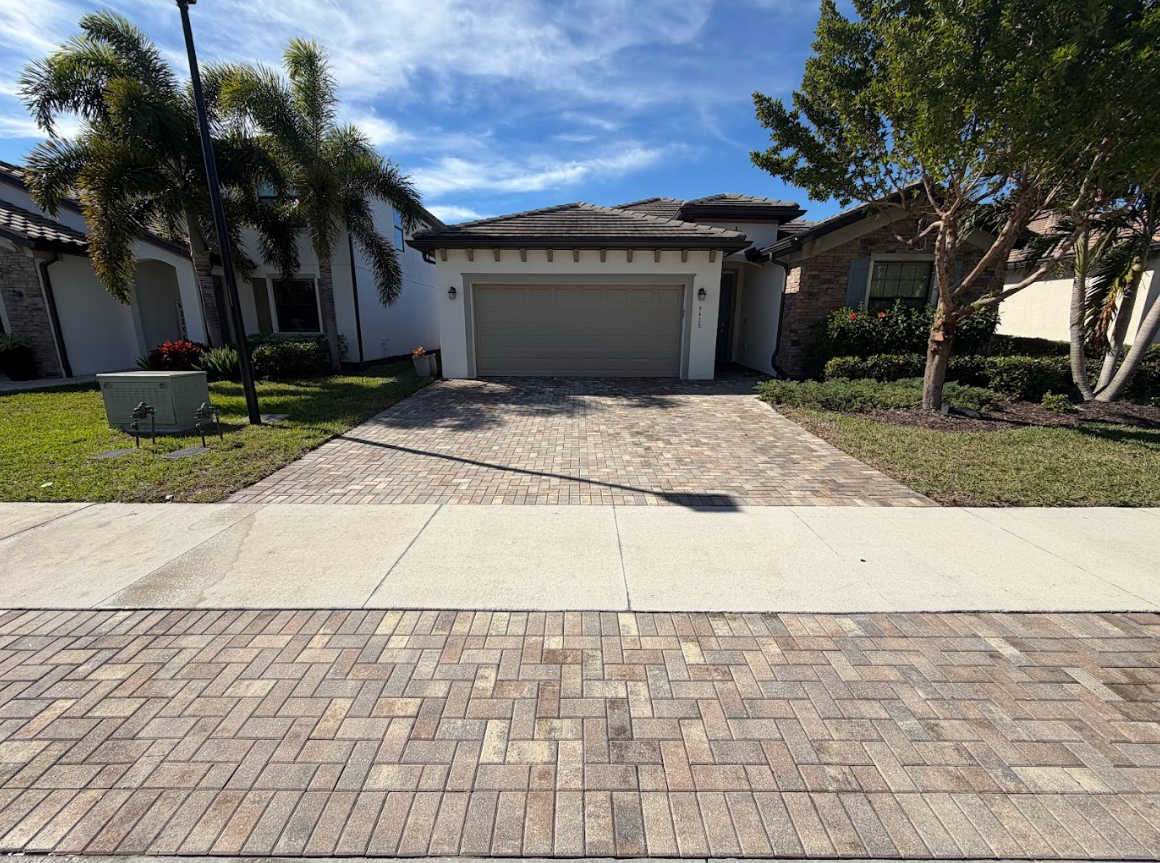 Suburban house with a garage and brick driveway, framed by palm trees under a clear sky