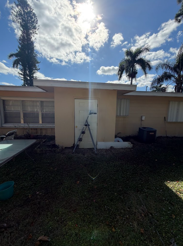 House exterior with ladder against a beige wall, sunny sky, and palm trees in the yard