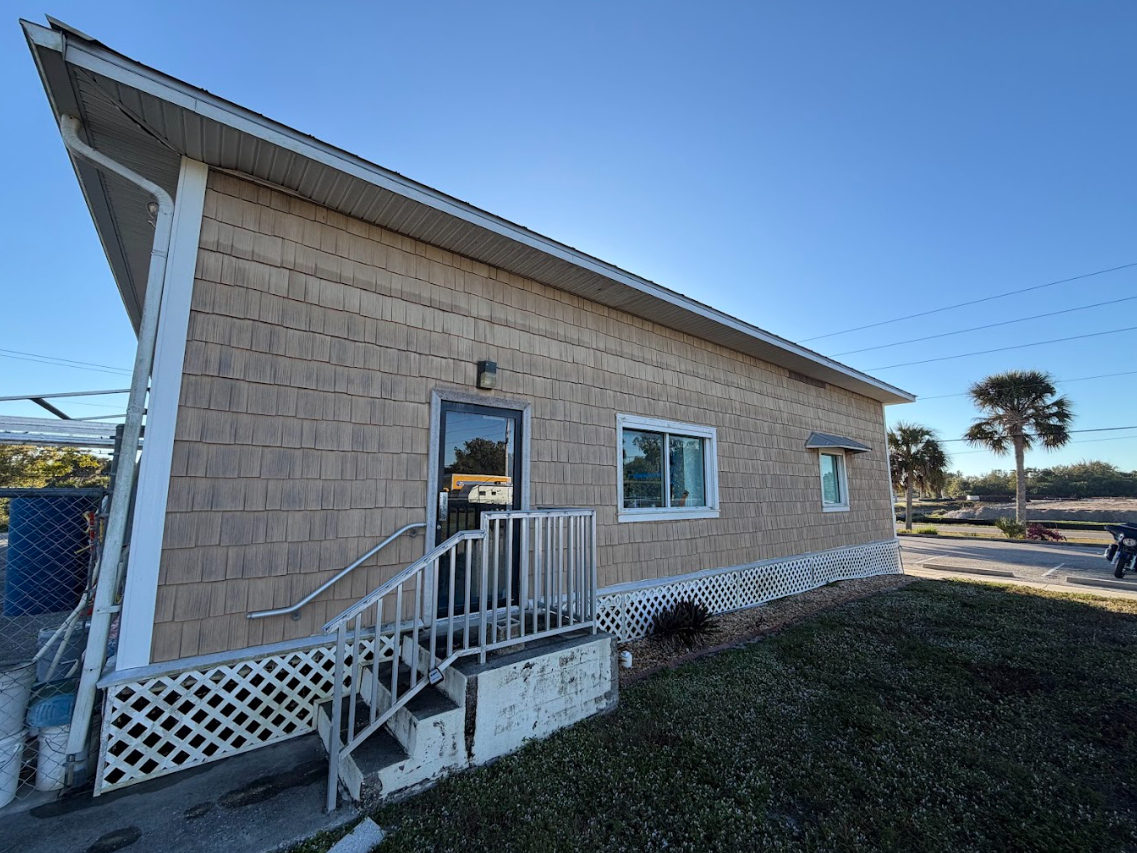 Small tan house with a sloped roof, front porch railing, and a window on a sunny day.