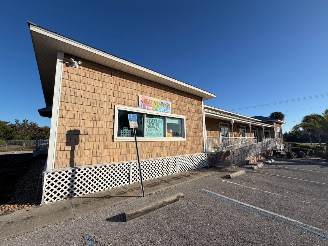 Small tan building with a slanted roof, front window sign, and empty parking lot under a blue sky
