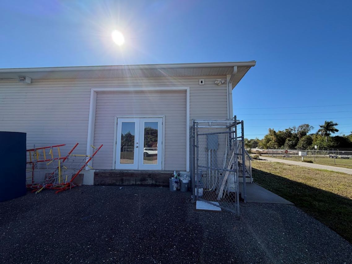 Exterior of a beige building with a white door, gravel ground, and metal cage by the wall under a bright sun