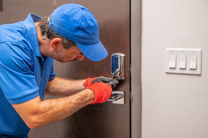 A man in a blue shirt and hat is fixing a door lock. A man in a blue shirt and hat is fixing a door lock.