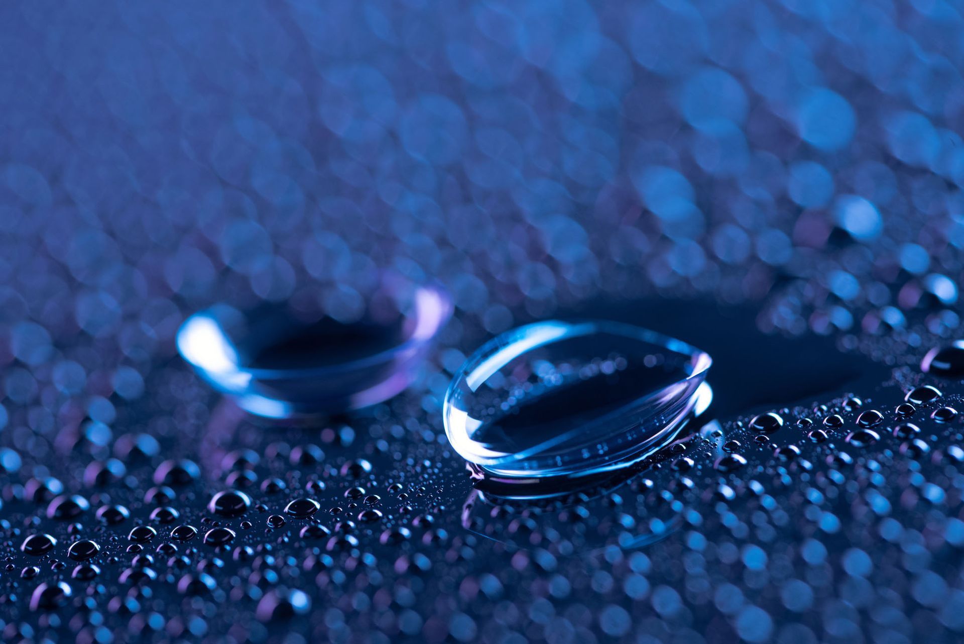 Due gocce d'acqua trasparenti su una superficie blu scuro e ruvida, illuminate da riflessi blu freddi.