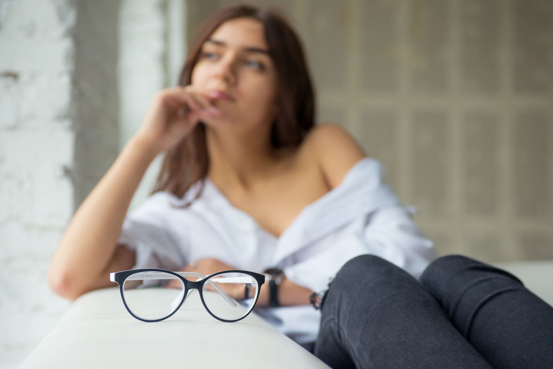 In primo piano, una donna sdraiata su un divano con gli occhiali indossa una camicia bianca e jeans scuri.