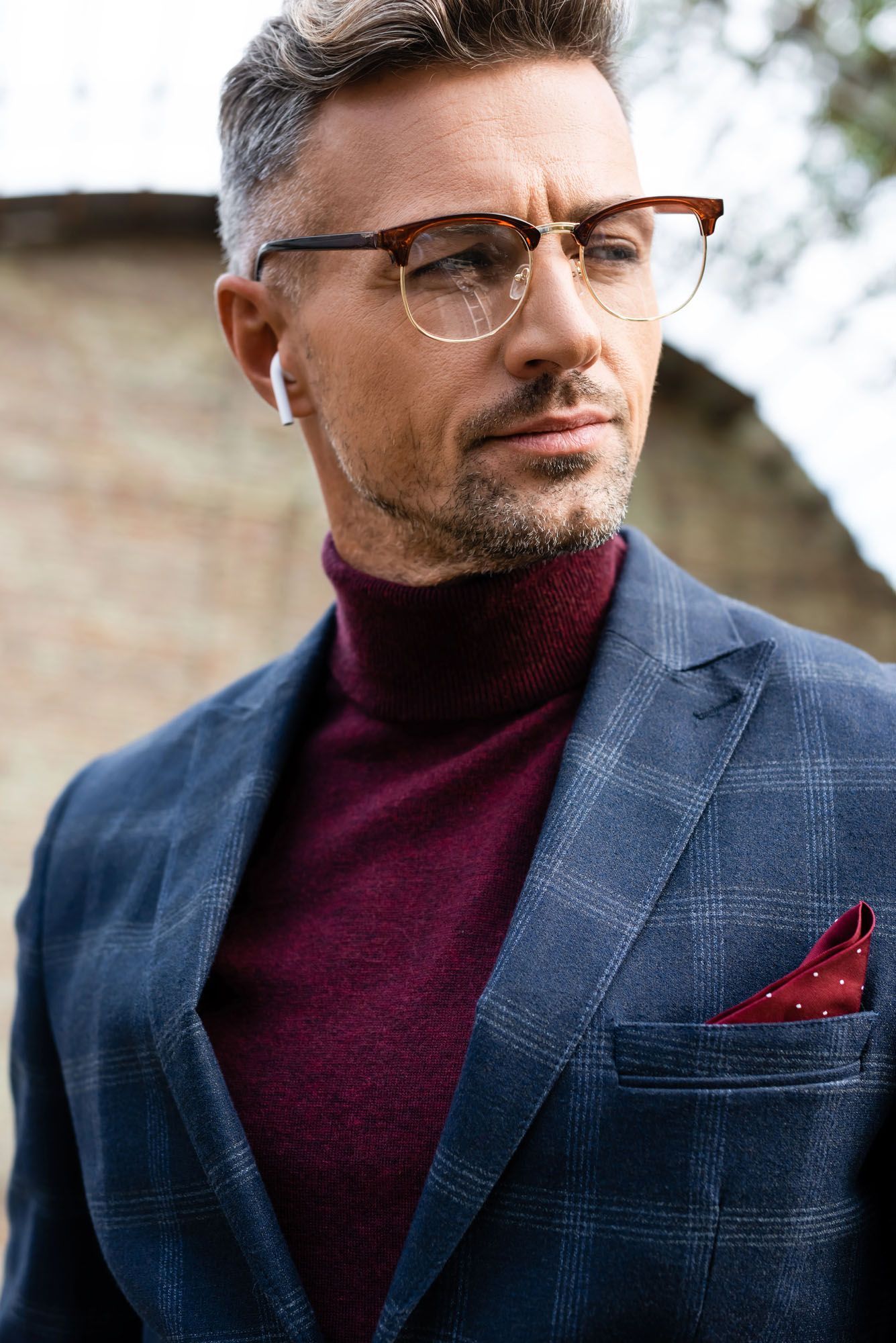 Uomo con gli occhiali, che indossa un blazer blu scuro e un dolcevita bordeaux, in piedi all'aperto con un fazzoletto da taschino piegato.