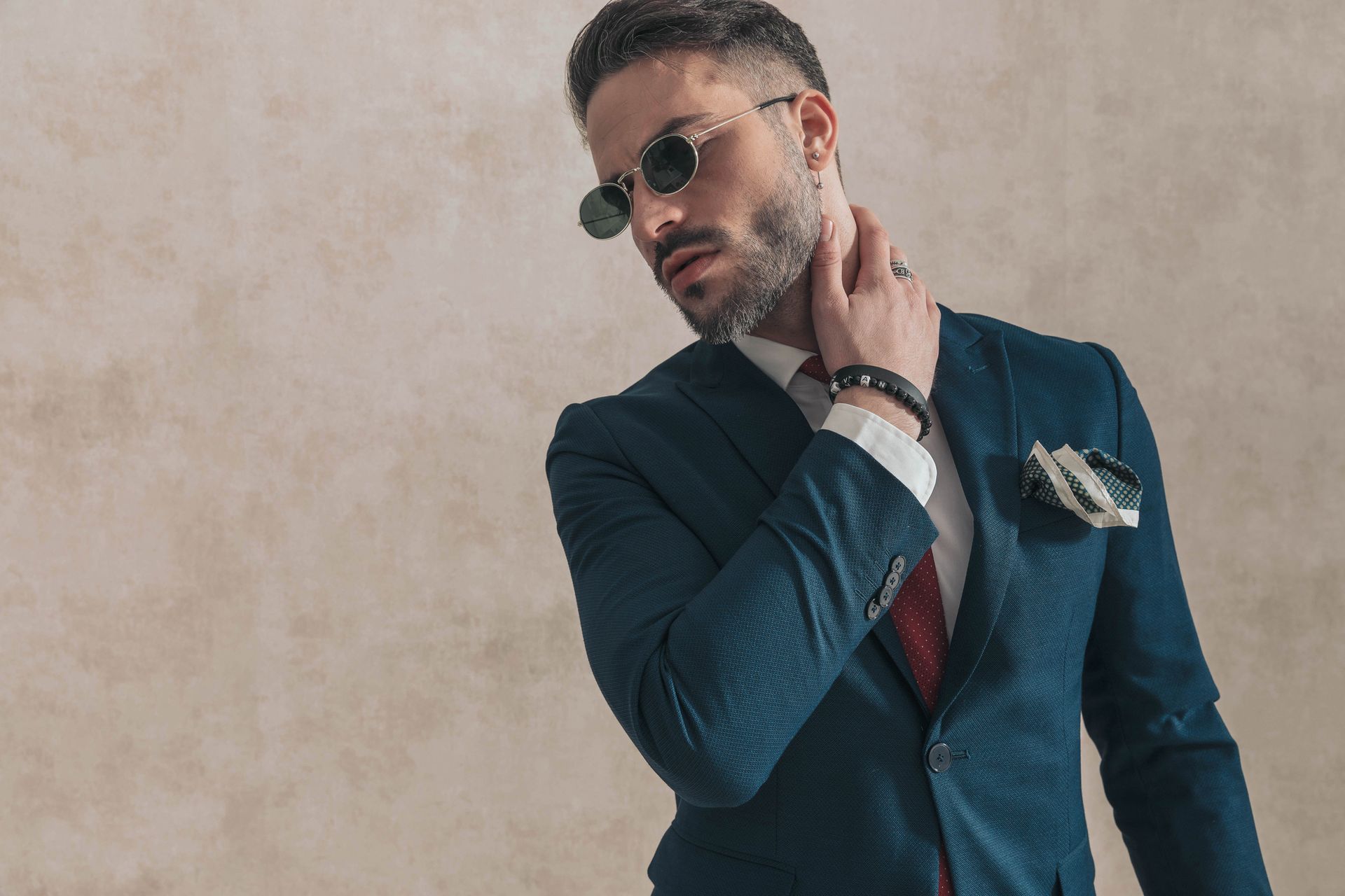 Un uomo in abito blu scuro e occhiali da sole posa contro un muro beige, con la mano sul collo.
