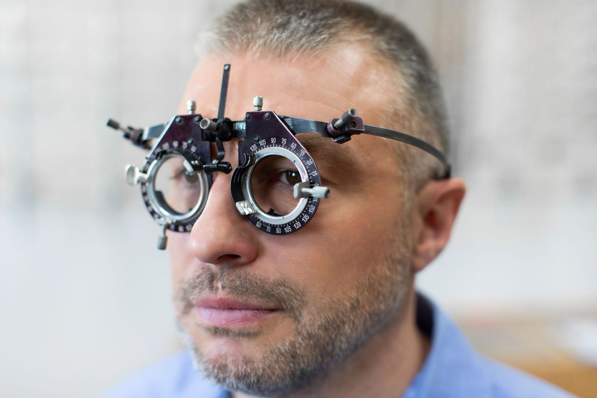 Uomo con occhiali di prova per una visita oculistica, visto di profilo su uno sfondo neutro.