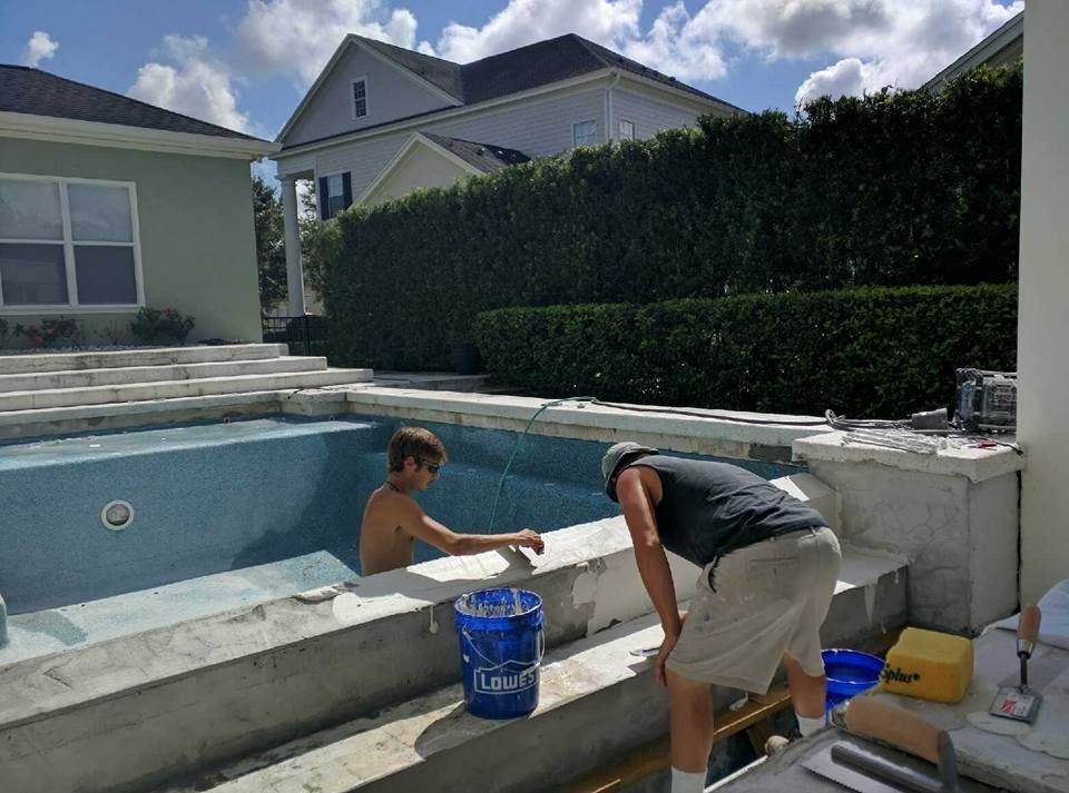 Two people working on a swimming pool: one in the pool, the other outside.