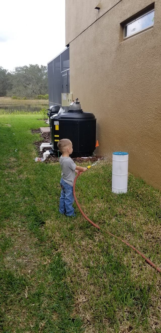 A young child holding a hose, spraying water on the grass next to a building and water tanks.