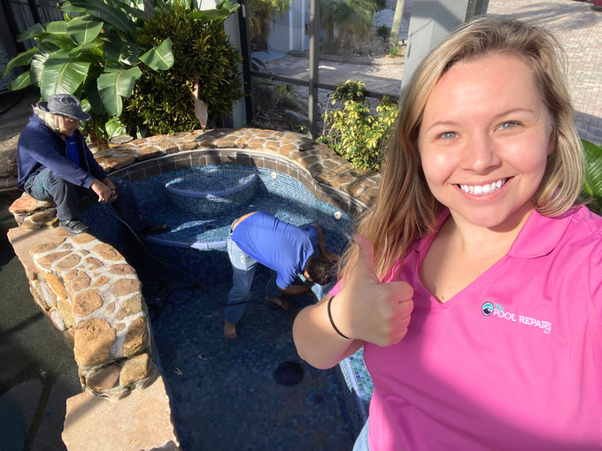 Smiling woman in pink shirt taking a selfie beside two workers cleaning a backyard pool