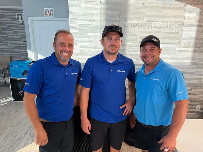 Three men in blue polo shirts and caps standing together indoors, smiling at the camera.
