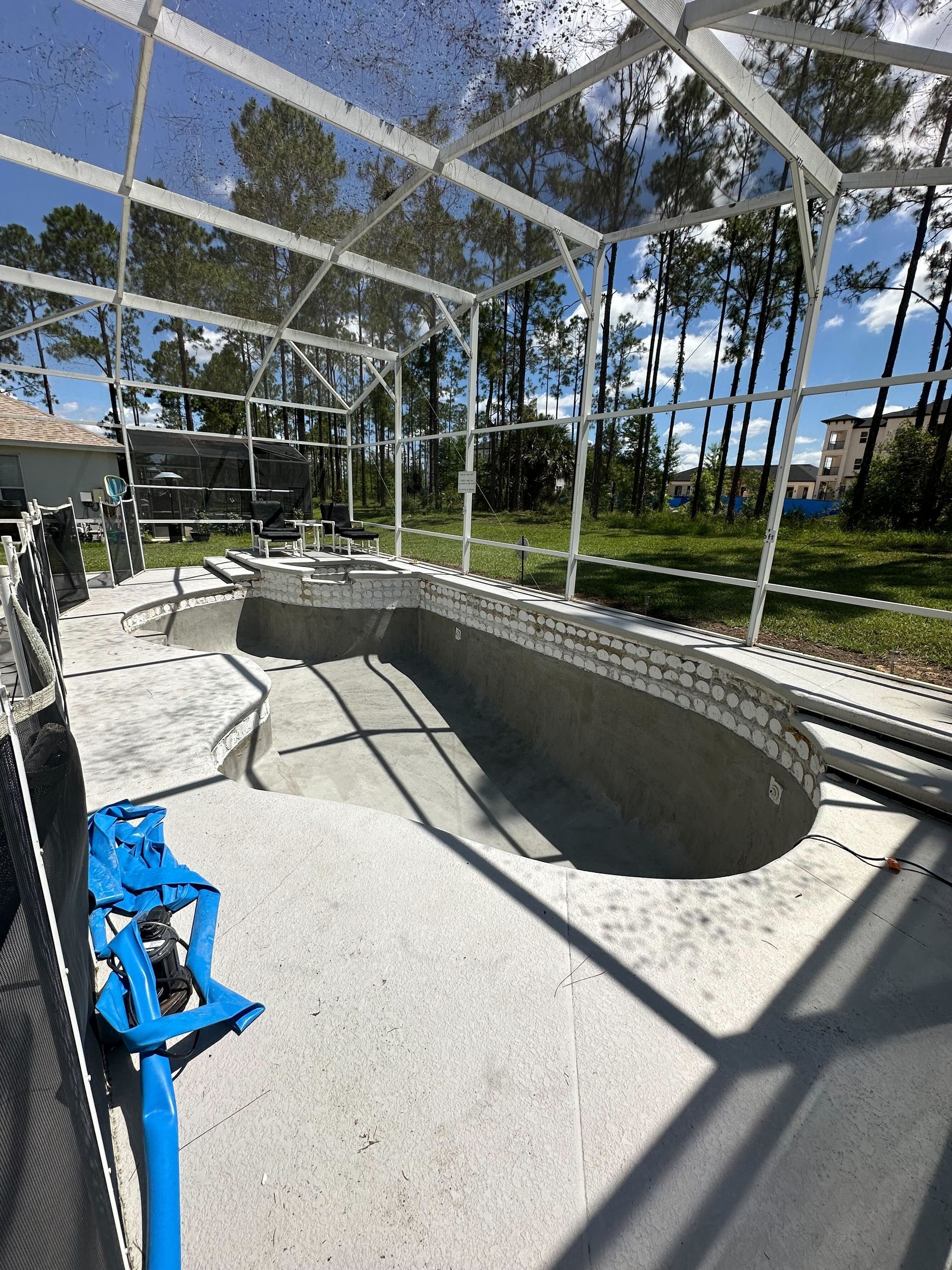 Empty, unfinished swimming pool inside a screened enclosure; concrete deck, trees in background.
