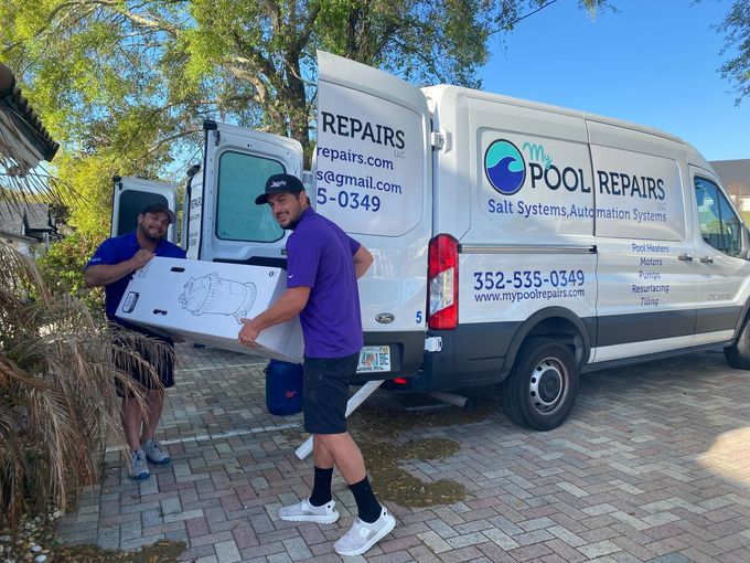 Two workers by a white Pool Repairs van, holding a box on a sunny driveway.