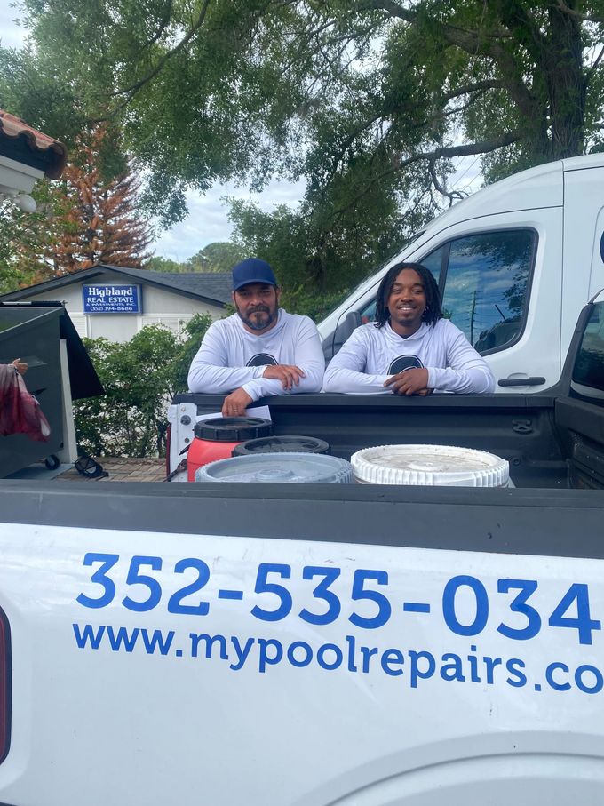 Two people behind a truck with pool service signage and phone number outdoors under trees