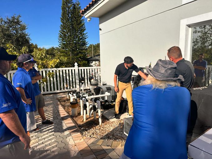 Group of people gathered around pool equipment outside. One man is pointing.
