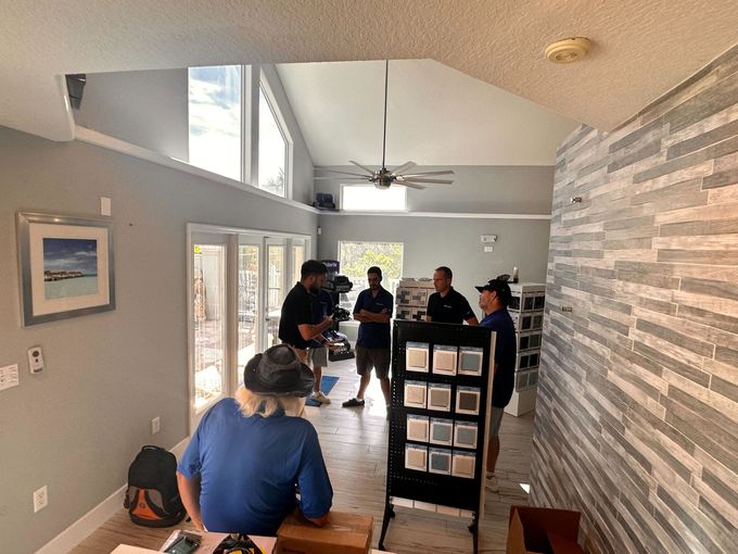 Group of people gathered indoors, looking at samples. Interior has grey walls, windows, and textured accent wall.