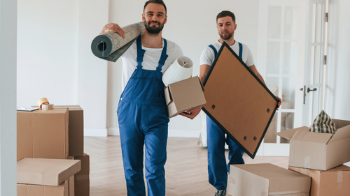 Two movers in blue overalls carrying boxes and wrapped furniture in a bright room