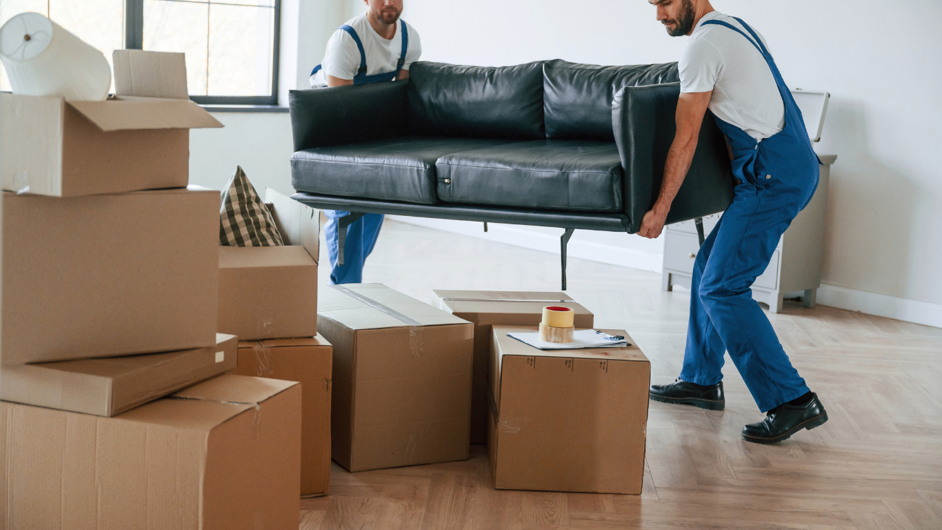 Two movers carrying a black couch through a room with moving boxes and packing supplies