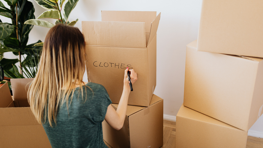 Person writing on a cardboard box among stacked moving boxes in a room with a plant