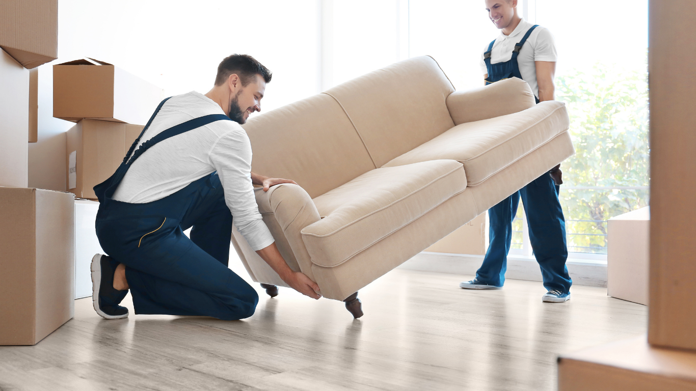Two movers carrying a beige sofa through a bright room with moving boxes.