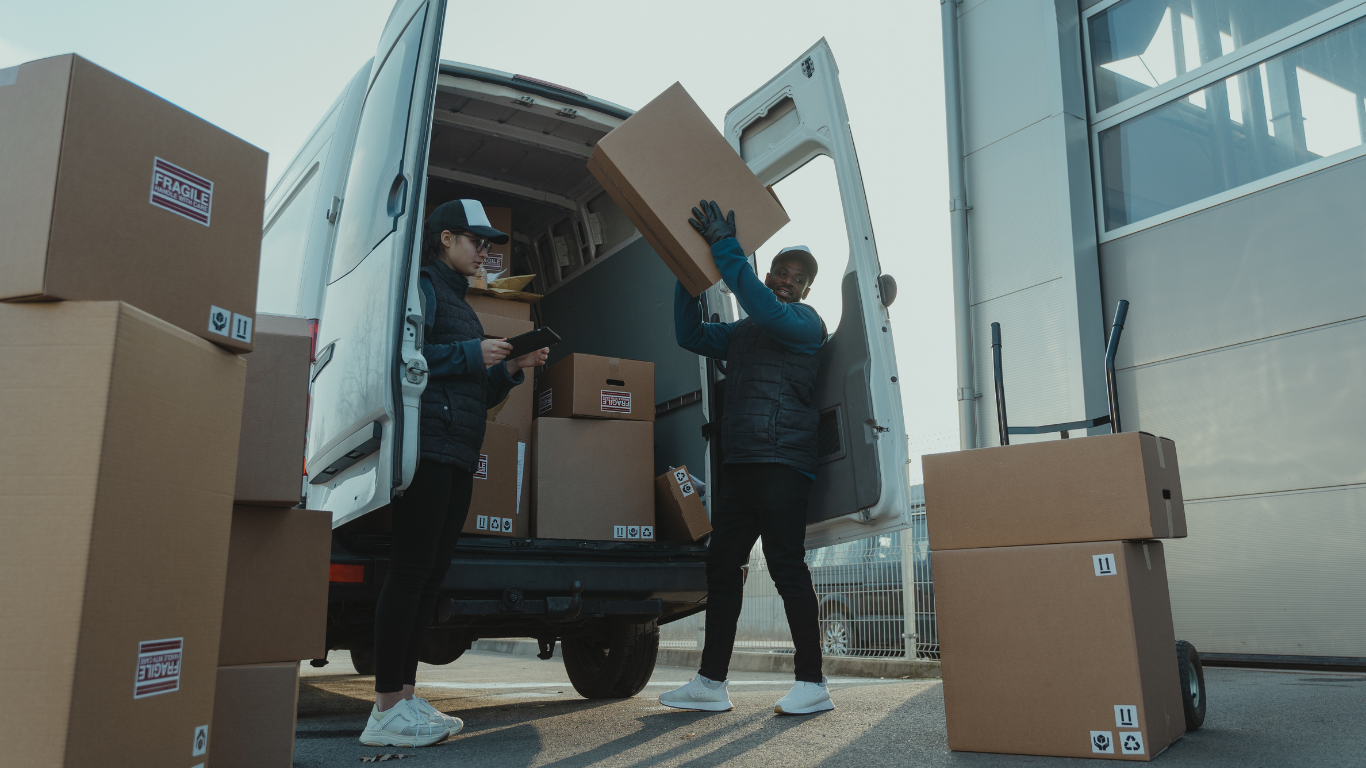 Two movers loading cardboard boxes into a white delivery truck outside a warehouse