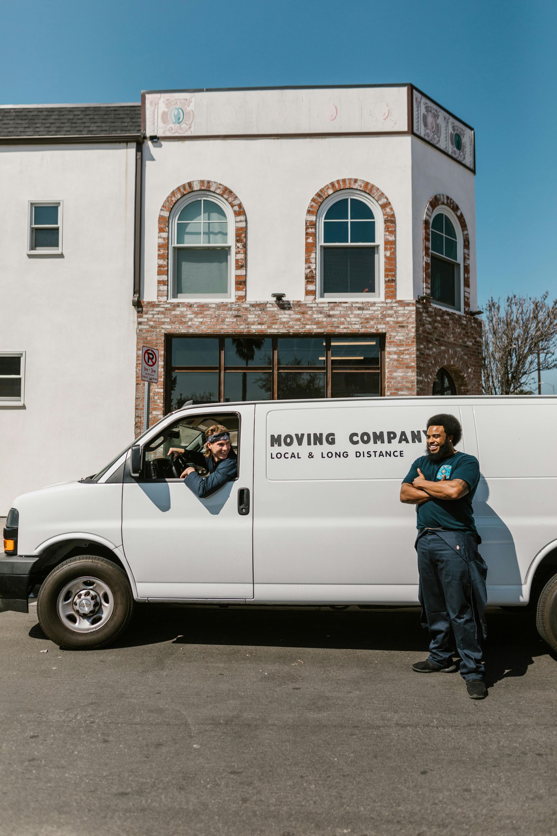 White work van parked in front of a stone storefront, with a person leaning against it under a clear blue sky.