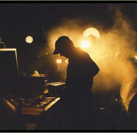 Silhouetted DJ wearing a cap at a smoky, golden-lit concert setup