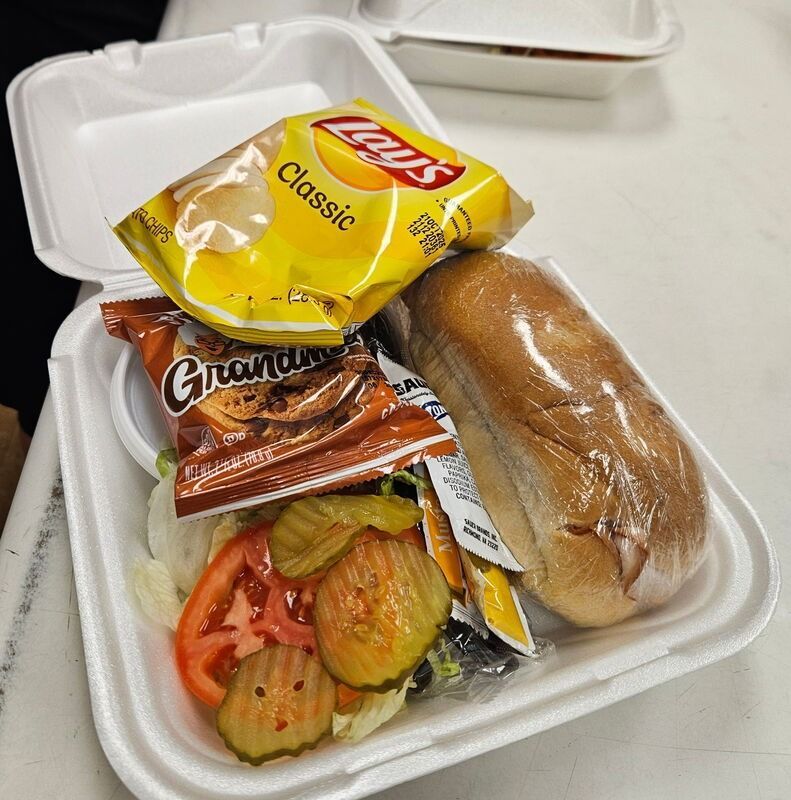 Takeout meal in a foam container with a sandwich, chips, roasted vegetables, and condiment packets