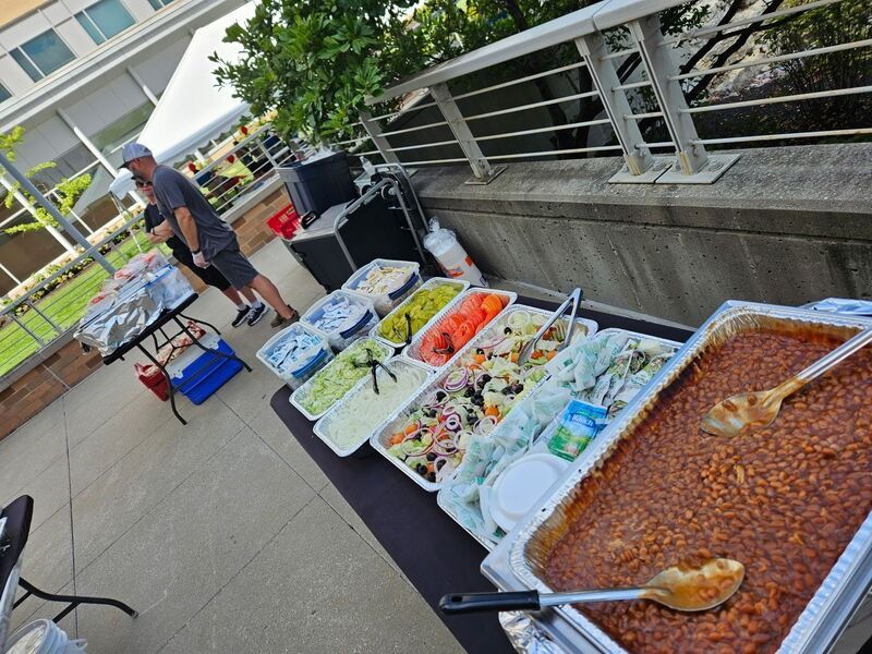 Outdoor buffet table with assorted salads, pasta, and a large baked dish beside a person serving food