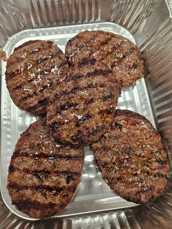 Four grilled burger patties in a foil tray, with dark char marks.