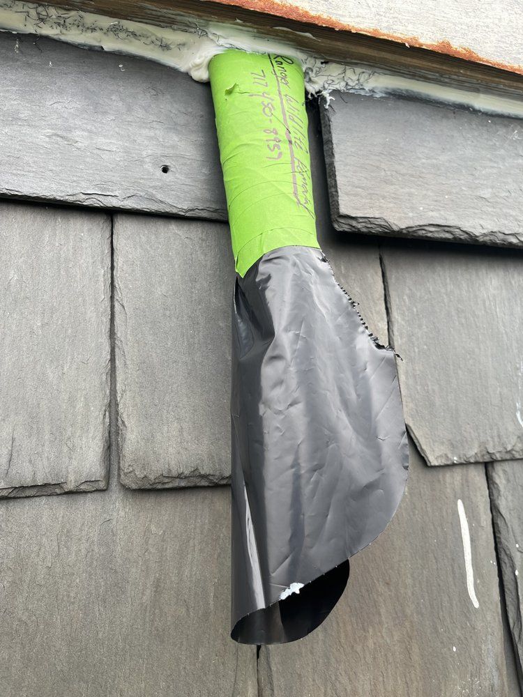 Green pipe wrapped in gray tape protruding from a gray shingled wall.